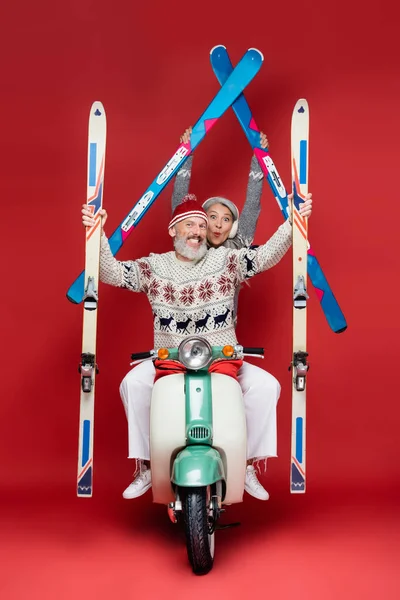 Happy interracial and middle aged skiers in sweaters holding skis while riding motor scooter on red — Stock Photo