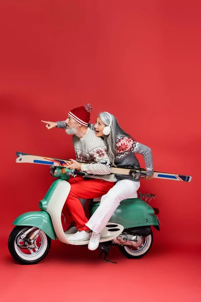 Surprised and interracial middle aged woman in sweater pointing with finger near husband holding skis while riding moped on red — Stock Photo