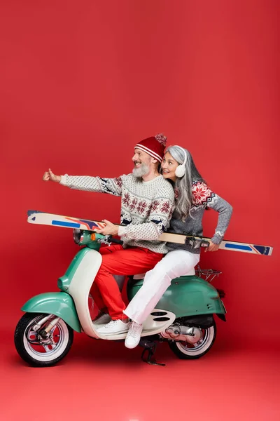 Smiling interracial and middle aged woman in sweater holding skis while husband showing thumb up and riding moped on red — Stock Photo