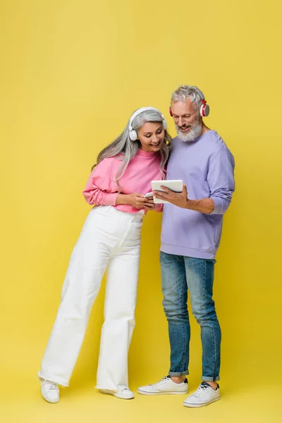 Comprimento total de feliz casal de meia-idade e interracial em fones de ouvido sem fio usando gadgets em amarelo — Fotografia de Stock