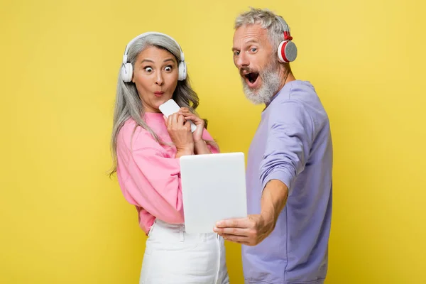 Sorprendida pareja de mediana edad e interracial en auriculares inalámbricos mirando tableta digital aislada en amarillo - foto de stock