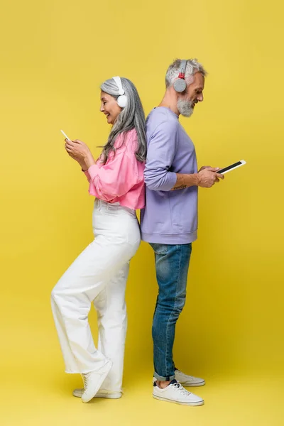 Side view of cheerful middle aged and interracial couple in wireless headphones using gadgets while standing back to back on yellow — Stock Photo