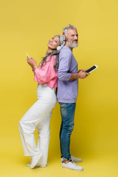 Longitud completa de feliz y interracial pareja de mediana edad en auriculares inalámbricos utilizando gadgets, mientras que de pie espalda con espalda en amarillo - foto de stock