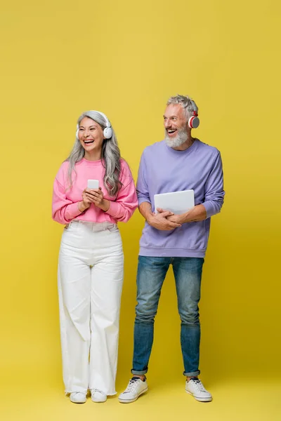 Comprimento total de feliz casal de meia idade e interracial em fones de ouvido sem fio segurando gadgets no amarelo — Fotografia de Stock