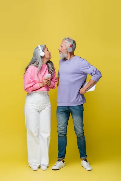 Comprimento total de engraçado interracial e meia idade casal em fones de ouvido sem fio segurando gadgets e olhando um para o outro em amarelo — Fotografia de Stock