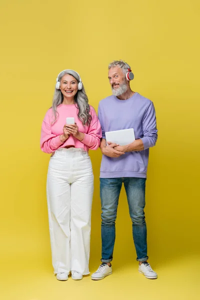 Comprimento total de casal multiétnico e sorrindo meia-idade em fones de ouvido sem fio segurando gadgets no amarelo — Fotografia de Stock