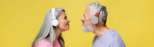 Vista lateral de la divertida pareja multiétnica y de mediana edad en auriculares inalámbricos que se miran aislados en amarillo, pancarta - foto de stock