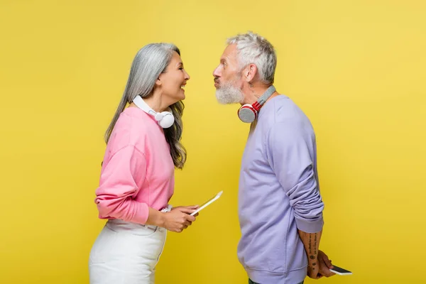 Seitenansicht eines lustigen multiethnischen Paares mittleren Alters mit drahtlosen Kopfhörern, die einander anschauen und isoliert auf gelb grimassieren — Stockfoto