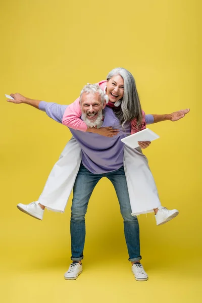 Cheerful middle aged man piggybacking happy asian wife with digital tablet on yellow — Stock Photo