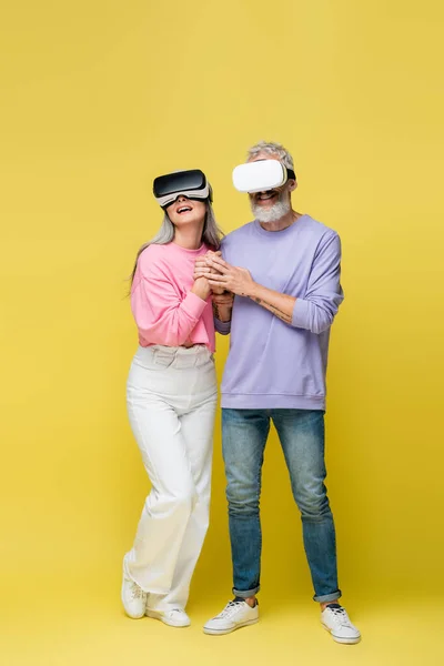 Full length of multiethnic and middle aged couple in vr headsets holding hands while gaming on yellow — Stock Photo