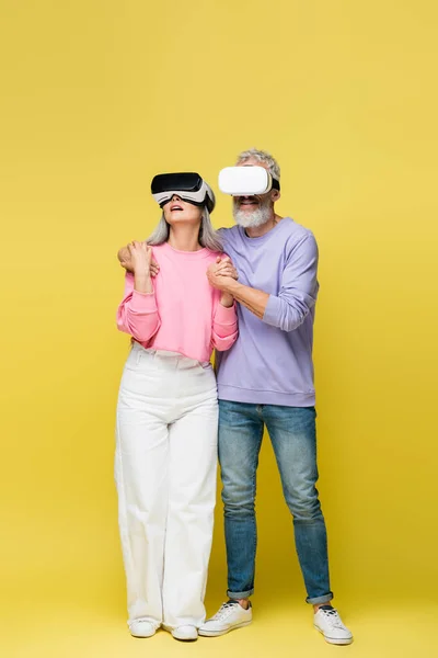 Full length of interracial and middle aged couple in vr headsets holding hands while gaming on yellow — Stock Photo