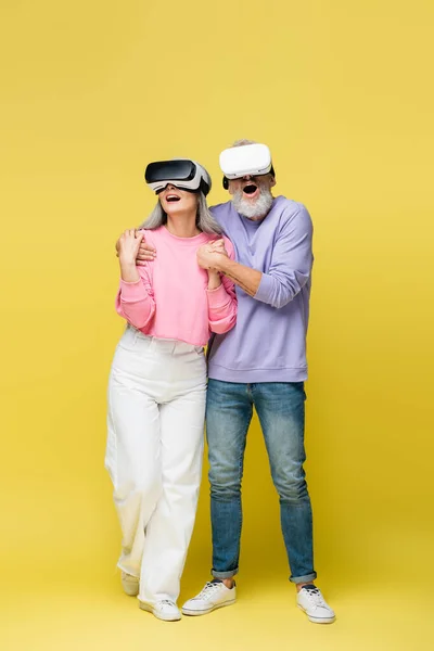 Full length of interracial and surprised middle aged couple in vr headsets holding hands while gaming on yellow — Stock Photo