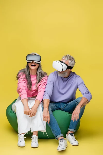 Interracial and happy middle aged couple in vr headsets sitting in bean bag chair on yellow — Stock Photo