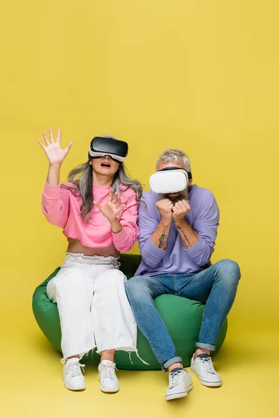 Interracial y asustado de mediana edad pareja en vr auriculares sentado en la silla de la bolsa de frijol, mientras que los juegos en amarillo — Stock Photo