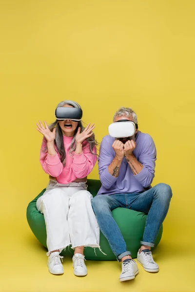 Interracial and afraid middle aged couple in vr headsets sitting in bean bag chair while gaming on yellow — Stock Photo