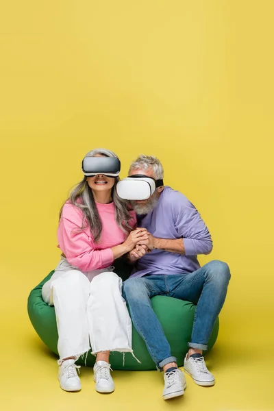 Interracial and scared mature couple in vr headsets holding hands and sitting in bean bag chair while gaming on yellow — Stock Photo