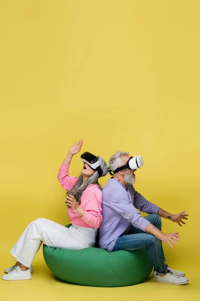Interracial e surpreso casal de meia idade em fones de ouvido vr sentado na cadeira saco feijão enquanto joga no amarelo — Fotografia de Stock