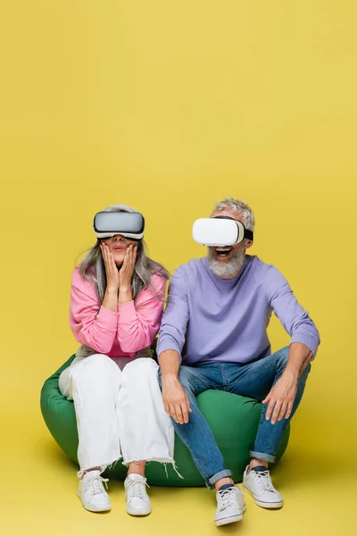 Interracial middle aged couple in vr headsets sitting in bean bag chair while gaming on yellow — Stock Photo