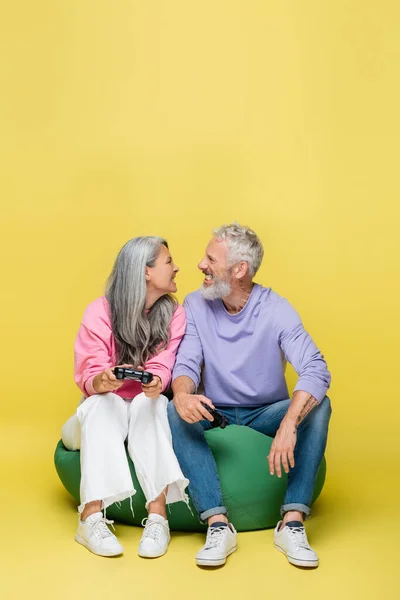 QUIIV, UCRÂNIA - AGOSTO 10, 2021: casal interracial e de meia idade segurando joysticks e dentes sorridentes enquanto sentado na cadeira saco feijão em amarelo — Fotografia de Stock