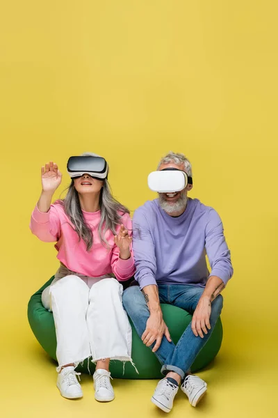 Interracial and middle aged couple in vr headsets sitting in bean bag chair and gaming on yellow — Stock Photo