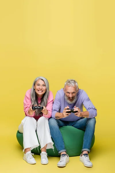 KYIV, UCRANIA - 10 de agosto de 2021: pareja interracial y excitada de mediana edad sosteniendo joysticks y jugando videojuegos en una silla de bolsa de frijoles en amarillo - foto de stock