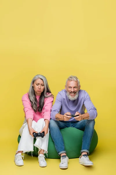 KIEW, UKRAINE - 10. AUGUST 2021: traurige asiatische Frau mittleren Alters hält Steuerknüppel in der Nähe eines glücklichen Ehemanns, der ein Videospiel auf gelb spielt — Stockfoto