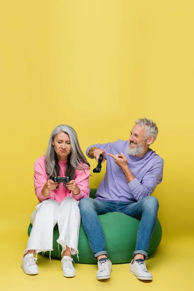 KYIV, UKRAINE - AUGUST 10, 2021: happy middle aged man pointing at sad asian wife losing video game on yellow — Stock Photo