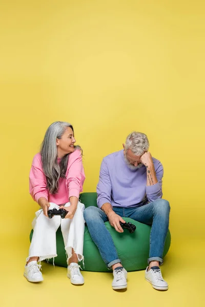 KYIV, UCRANIA - 10 de agosto de 2021: mujer de mediana edad asiática feliz sosteniendo el joystick cerca del triste esposo perdiendo el videojuego en amarillo - foto de stock