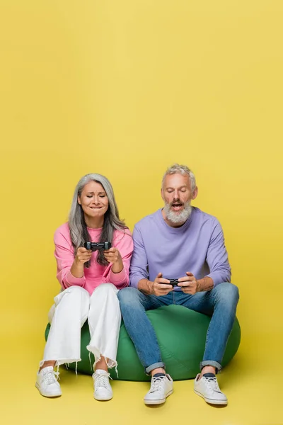 KYIV, UCRANIA - 10 de agosto de 2021: pareja interracial de mediana edad tensa sosteniendo joysticks y jugando videojuegos en amarillo - foto de stock