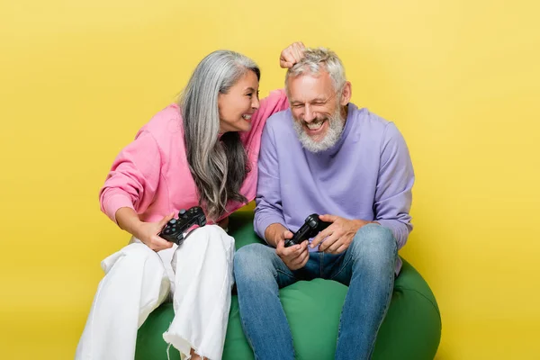 KYIV, UKRAINE - AUGUST 10, 2021: smiling asian woman knocking head of middle aged husband with joystick isolated on yellow — Stock Photo