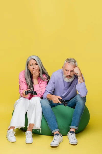 KYIV, UKRAINE - AUGUST 10, 2021: tired interracial middle aged couple holding joysticks and playing video game on yellow — Stock Photo