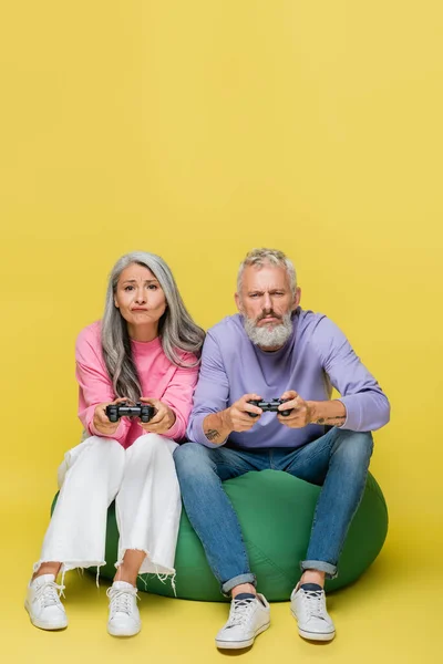 KYIV, UCRANIA - 10 de agosto de 2021: pareja interracial de mediana edad disgustada sosteniendo joysticks y jugando videojuegos en amarillo - foto de stock