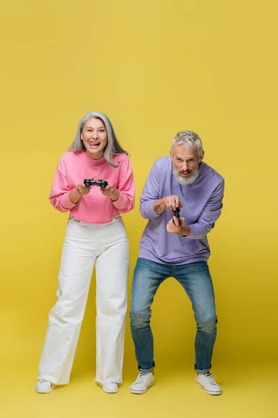 QUIIV, UCRÂNIA - 10 de agosto de 2021: comprimento total de casal feliz interracial e maduro jogando videogame em amarelo — Fotografia de Stock