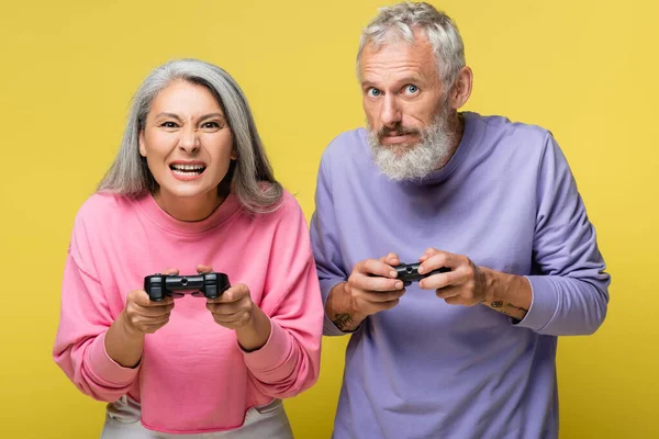 KYIV, UCRANIA - 10 de agosto de 2021: pareja interracial tensa y madura jugando videojuegos aislados en amarillo - foto de stock