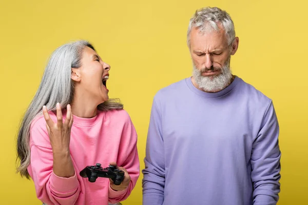 KYIV, UCRAINA - 10 AGOSTO 2021: donna asiatica urla e tiene il joystick vicino al triste marito maturo isolato sul giallo — Foto stock