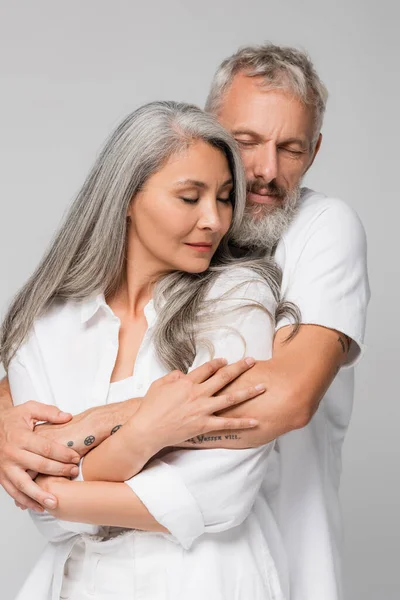 Pleased interracial and mature couple hugging isolated on grey — Stock Photo