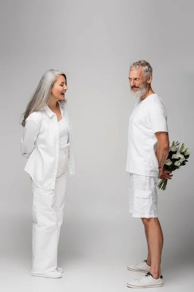 Full length of happy middle aged man hiding bouquet of flowers behind back near asian wife on grey — Stock Photo