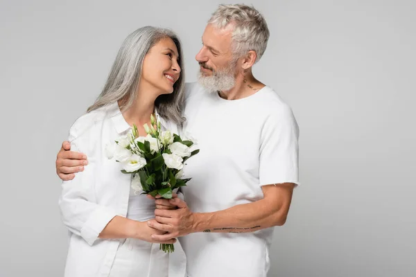 Happy interracial and mature couple holding flowers and looking at each other isolated on grey — Stock Photo