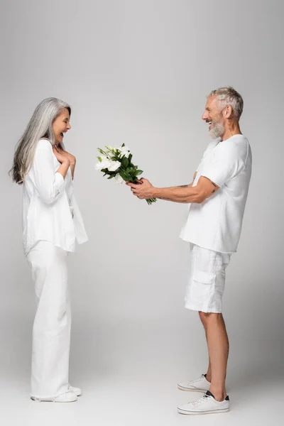 Vista lateral del hombre de mediana edad feliz dando ramo de flores a la esposa asiática asombrada en gris - foto de stock