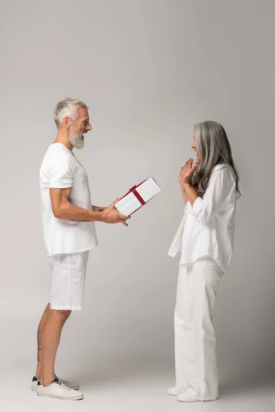 Повна довжина щасливого чоловіка середнього віку, що дарує загорнуту подарункову коробку, щоб здивувати азіатську дружину на сірому — стокове фото