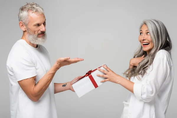 Happy middle aged man pointing with hand at amazed asian wife while giving wrapped gift box isolated on grey — Stock Photo