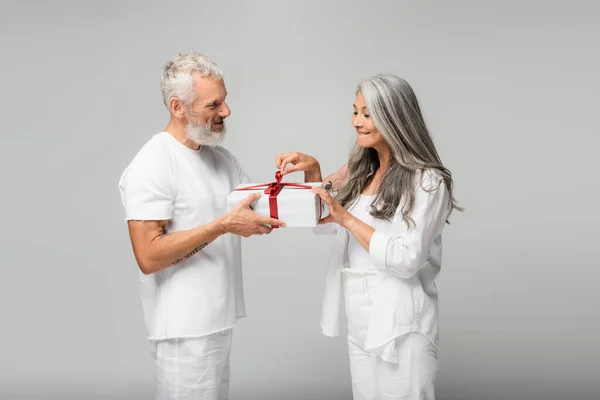 Happy middle aged man holding wrapped gift box near curious asian wife isolated on grey — Stock Photo