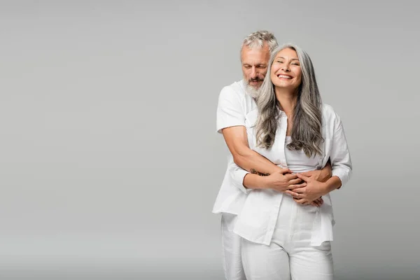 Happy mature man with closed eyes hugging pleased asian wife isolated on grey — Stock Photo