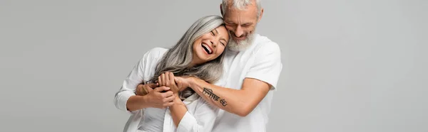 Happy mature man with closed eyes hugging joyful asian wife isolated on grey, banner — Stock Photo