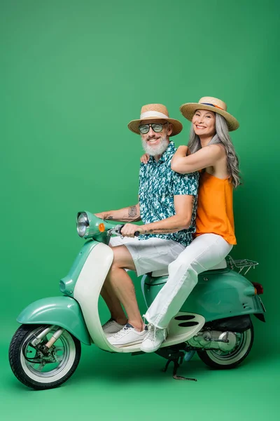 Mujer asiática feliz en sombrero de sol y marido de mediana edad a caballo moto en verde - foto de stock