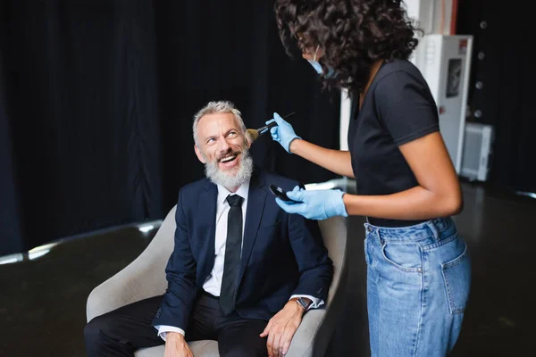 Maquilleur afro-américain en masque médical et gants en latex appliquant de la poudre visage sur homme d'affaires joyeux — Photo de stock