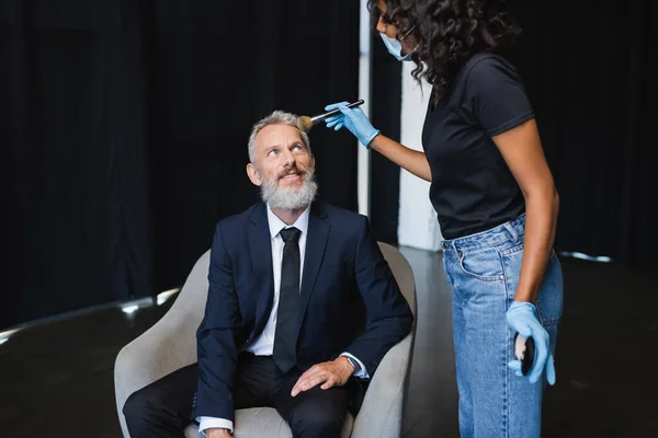 Curly african american makeup artist in medical mask and latex gloves applying face powder on smiling businessman — Stock Photo