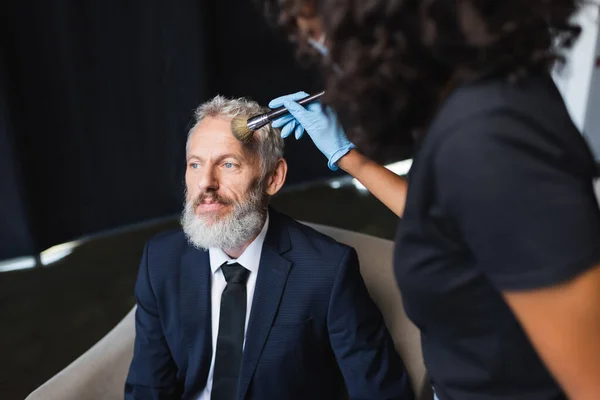 Blurred african american makeup artist in latex glove applying face powder on businessman — Stock Photo