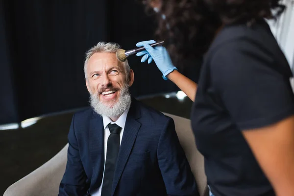 Blurred african american makeup artist in latex glove applying face powder on happy businessman — Stock Photo