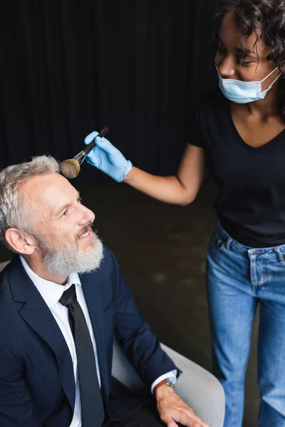 Artista de maquillaje afroamericano rizado en máscara médica y guante de látex aplicando polvo facial en hombre de negocios sonriente - foto de stock